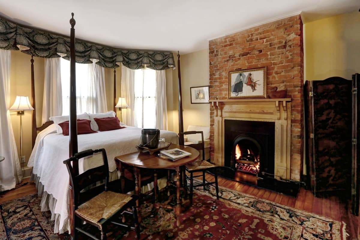 A four-poster bed with a fireplace, a dining table, and a patterned rug in a room with brick walls and wooden furniture.