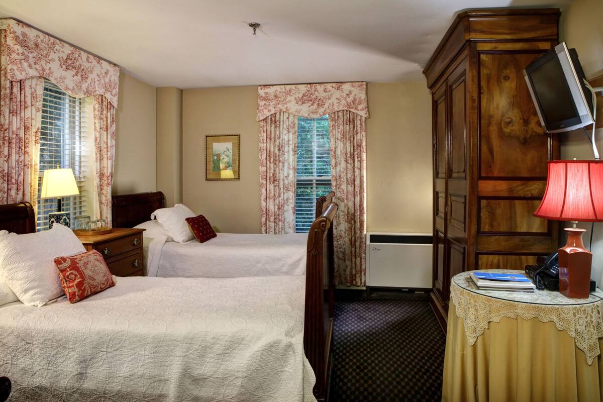 A hotel room with two beds, a wooden wardrobe, a television on a stand, and a small table with a lamp and a book.