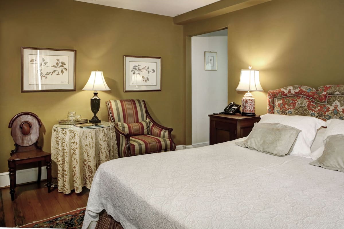 A hotel room with a large bed, two bedside tables with lamps, a striped armchair, a small round table with a lamp, and framed floral artwork on the walls.