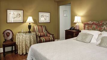 A hotel room with a large bed, two bedside tables with lamps, a striped armchair, a small round table with a lamp, and framed floral artwork on the walls.