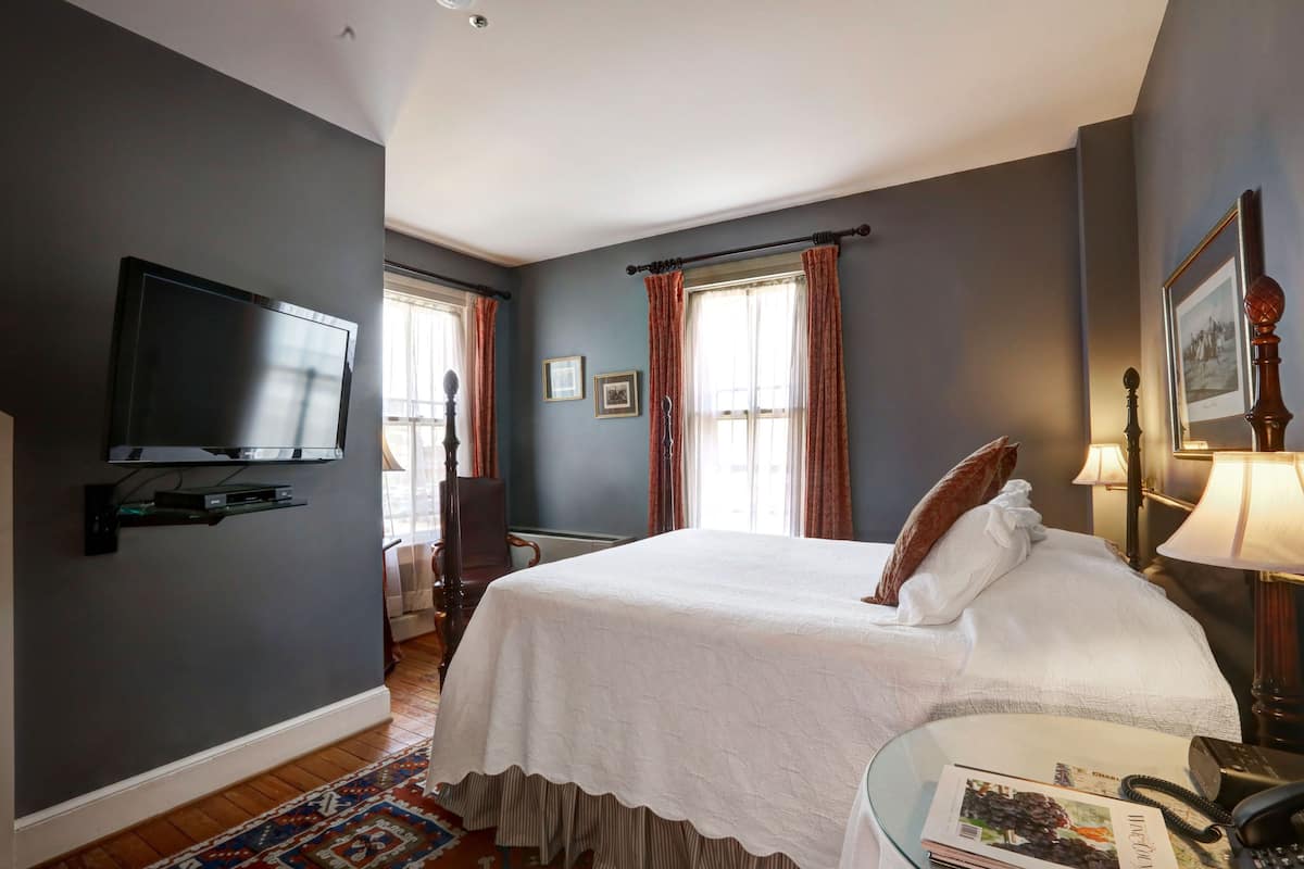 A hotel room with a large bed, a flat-screen TV mounted on the wall, a bedside table with a lamp, and a window with curtains.