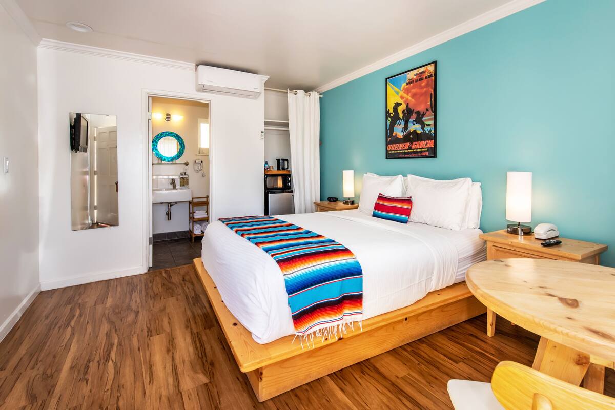 A hotel room with a bed, bedside tables, a small wooden table, a bathroom with a mirror, and a poster on the wall.