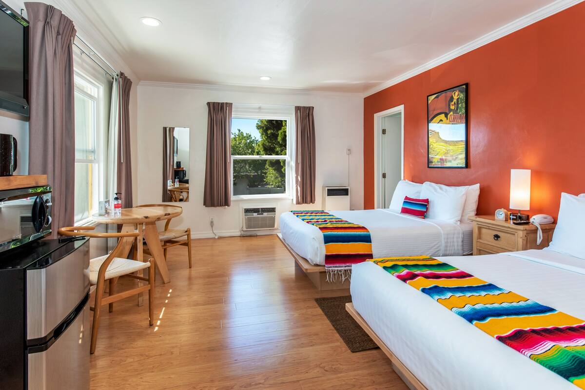 A hotel room with two beds, a dining area, a kitchen, and a large window.