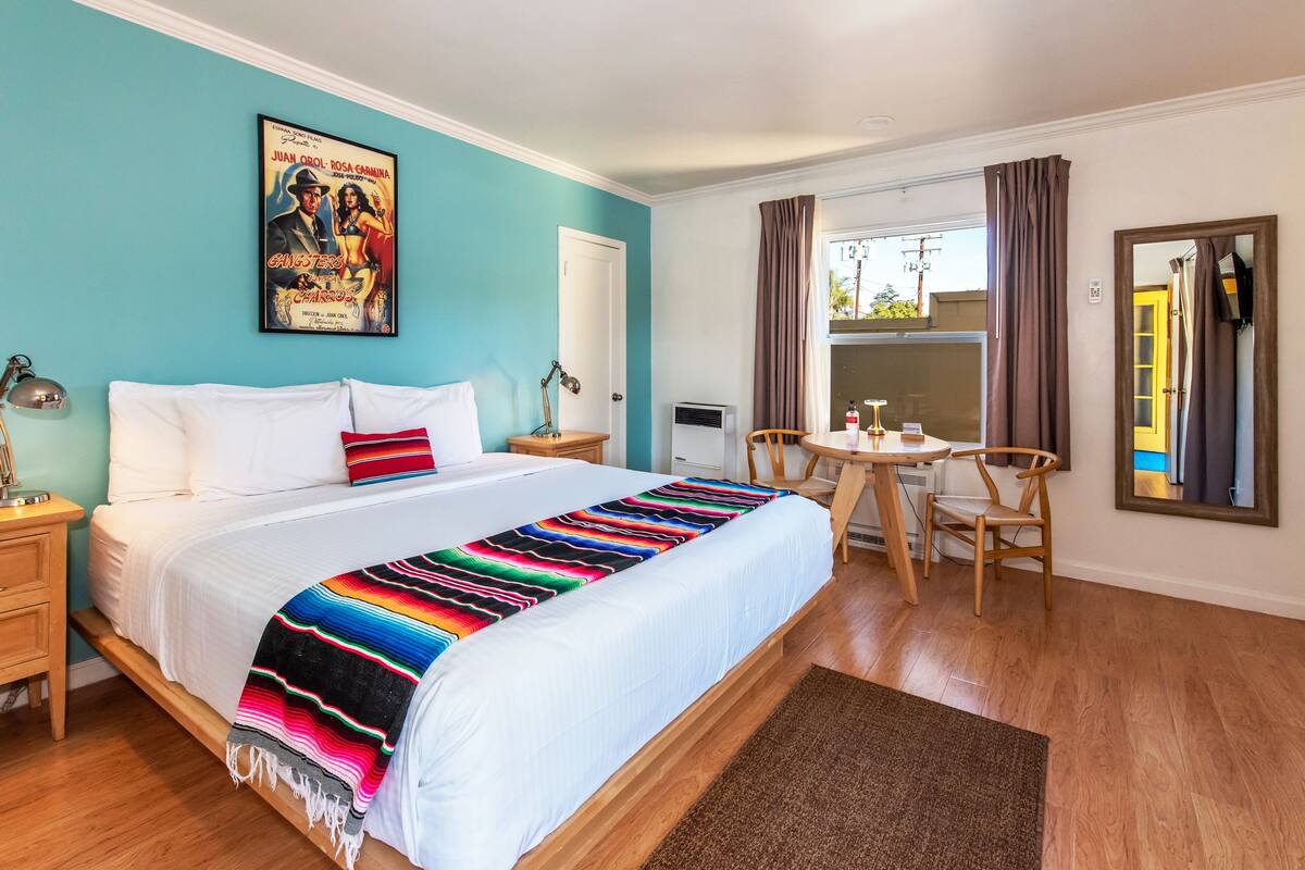 A hotel room with a bed, a desk, and a chair. There is a poster on the wall and a window with curtains.