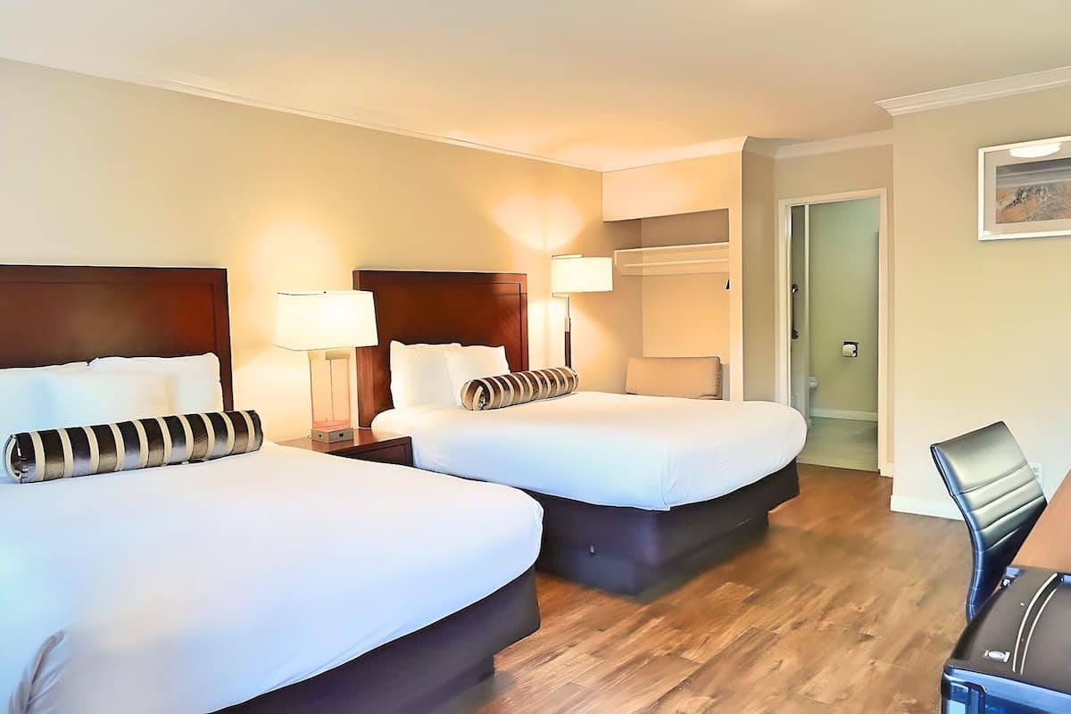 A hotel room with two beds, a wooden floor, a desk with a chair, and a door leading to a bathroom.
