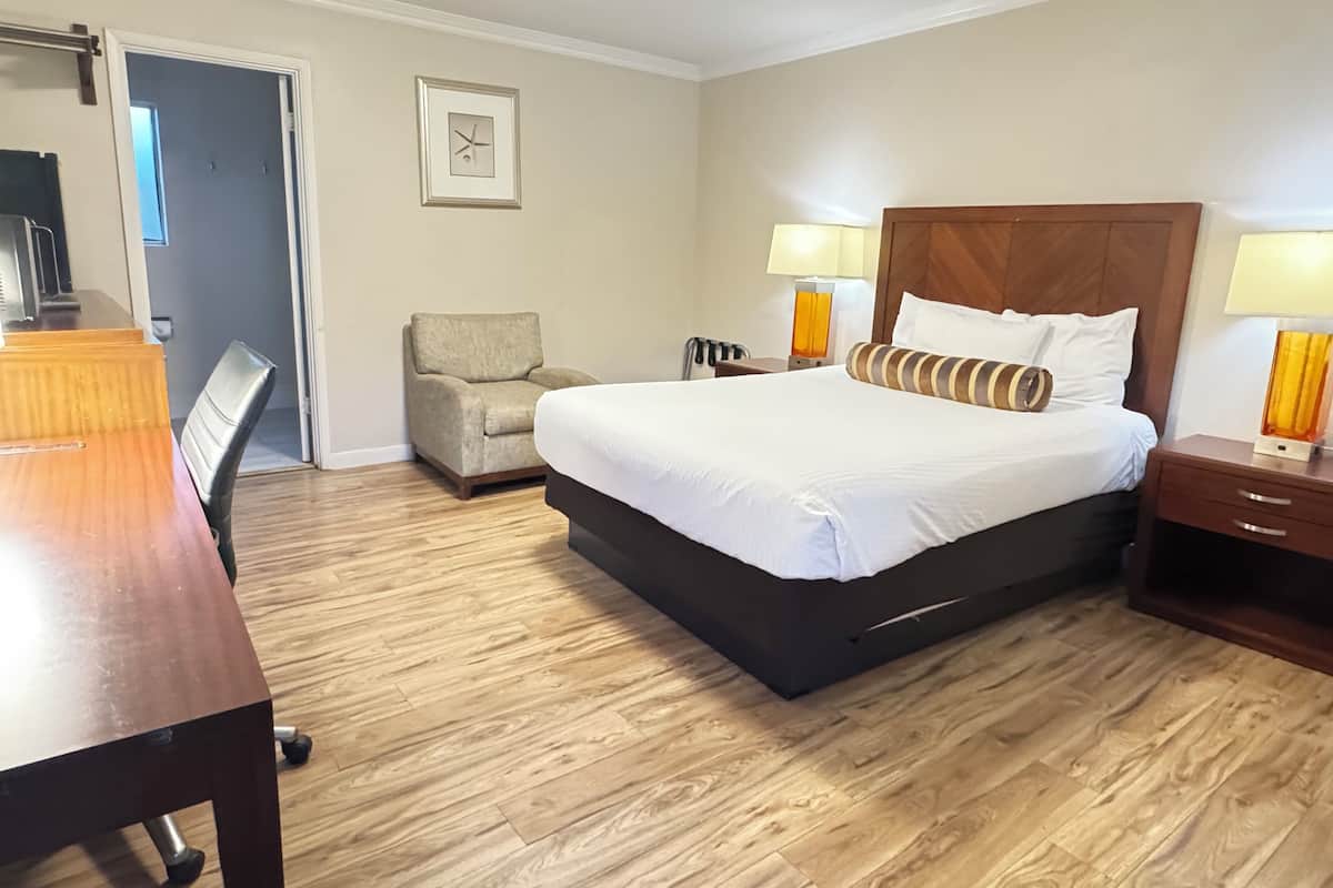 A hotel room with a large bed, two bedside tables with lamps, a chair, a desk, and a wooden floor.