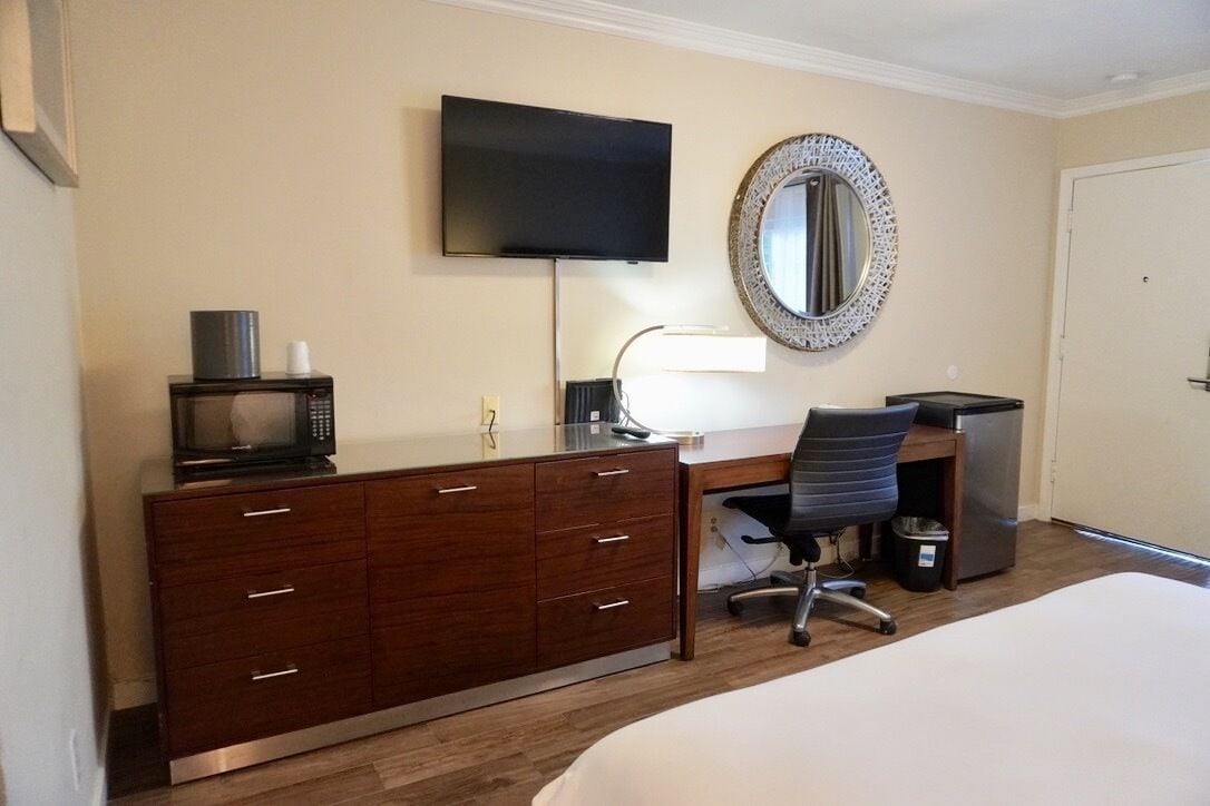 A hotel room with a bed, a desk with a computer, a chair, a television, a microwave, and a dresser.