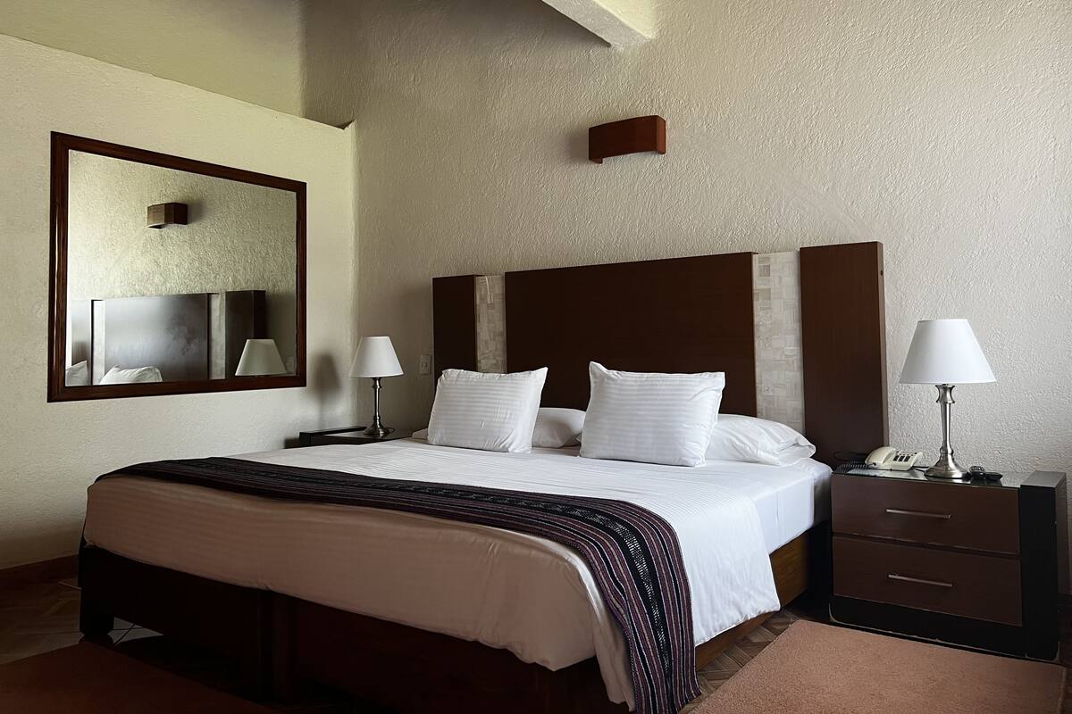 Habitación de hotel con una cama grande, dos mesitas de noche, un tocador, un espejo y un techo con vigas a la vista.