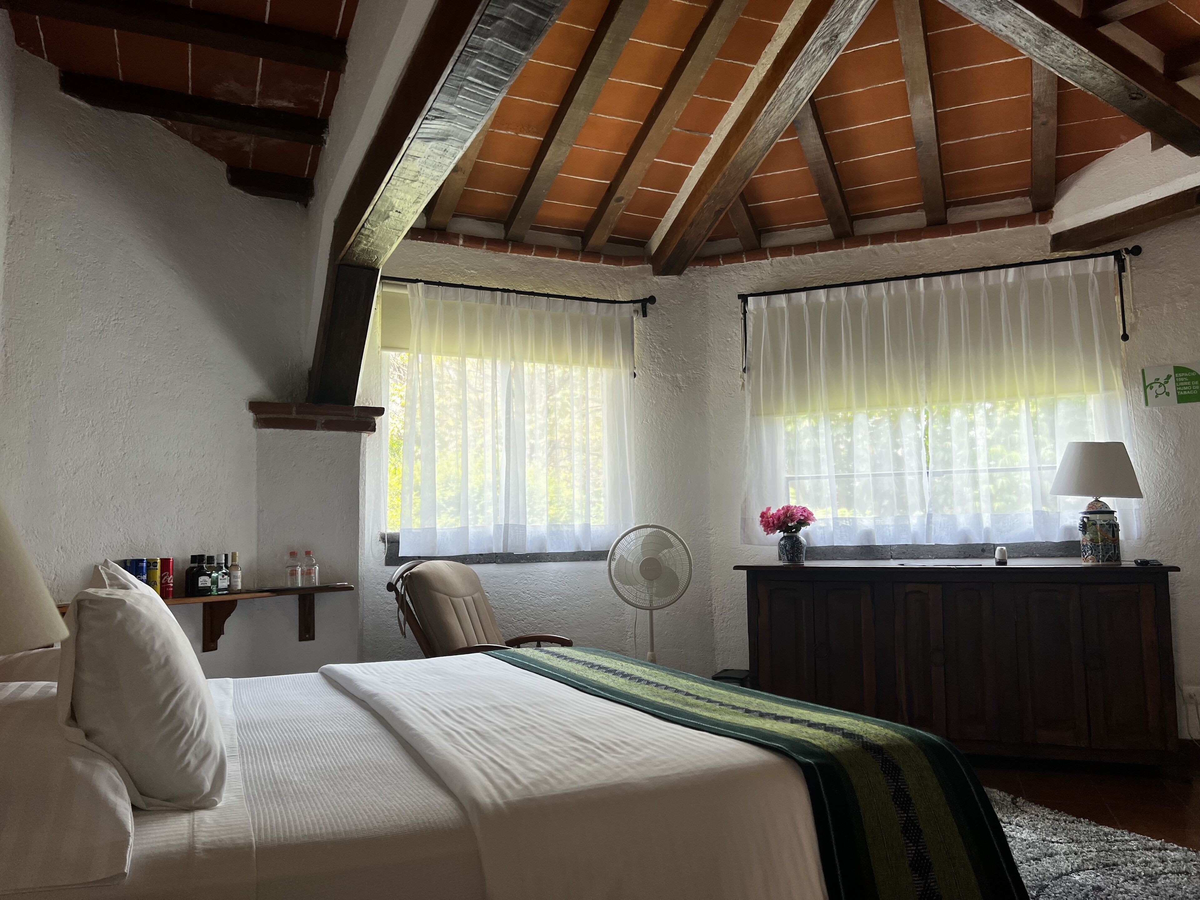 A bedroom with a bed, a chair, a bedside table with a lamp, a fan, and a window with sheer curtains.
