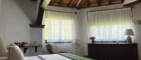 A bedroom with a bed, a chair, a bedside table with a lamp, a fan, and a window with sheer curtains.