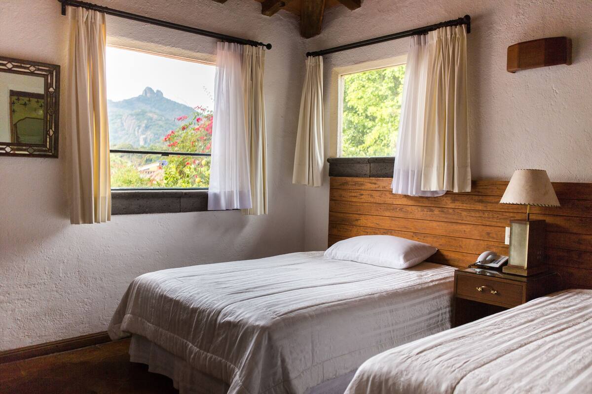 Un dormitorio con dos camas, cabeceros de madera, una mesita de noche con lámpara y una ventana con cortinas.