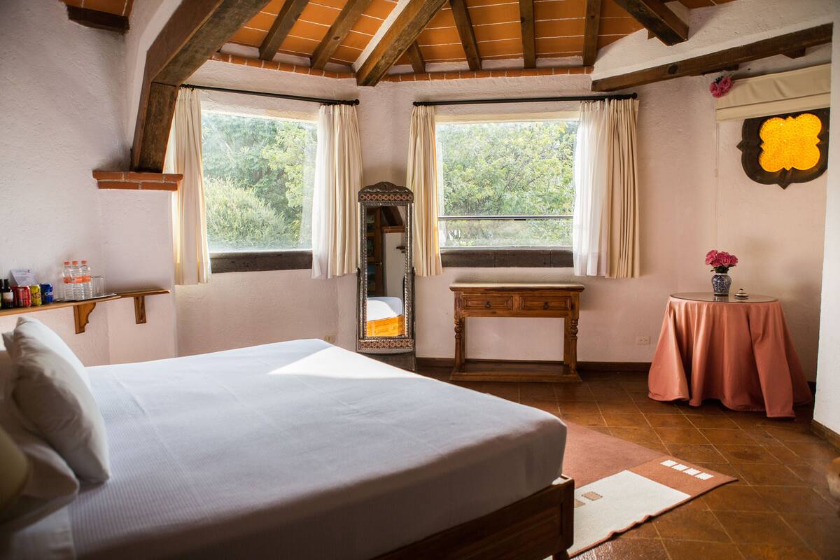 Un dormitorio con cama, cómoda, espejo, una mesita con un florero y ventana con cortinas.