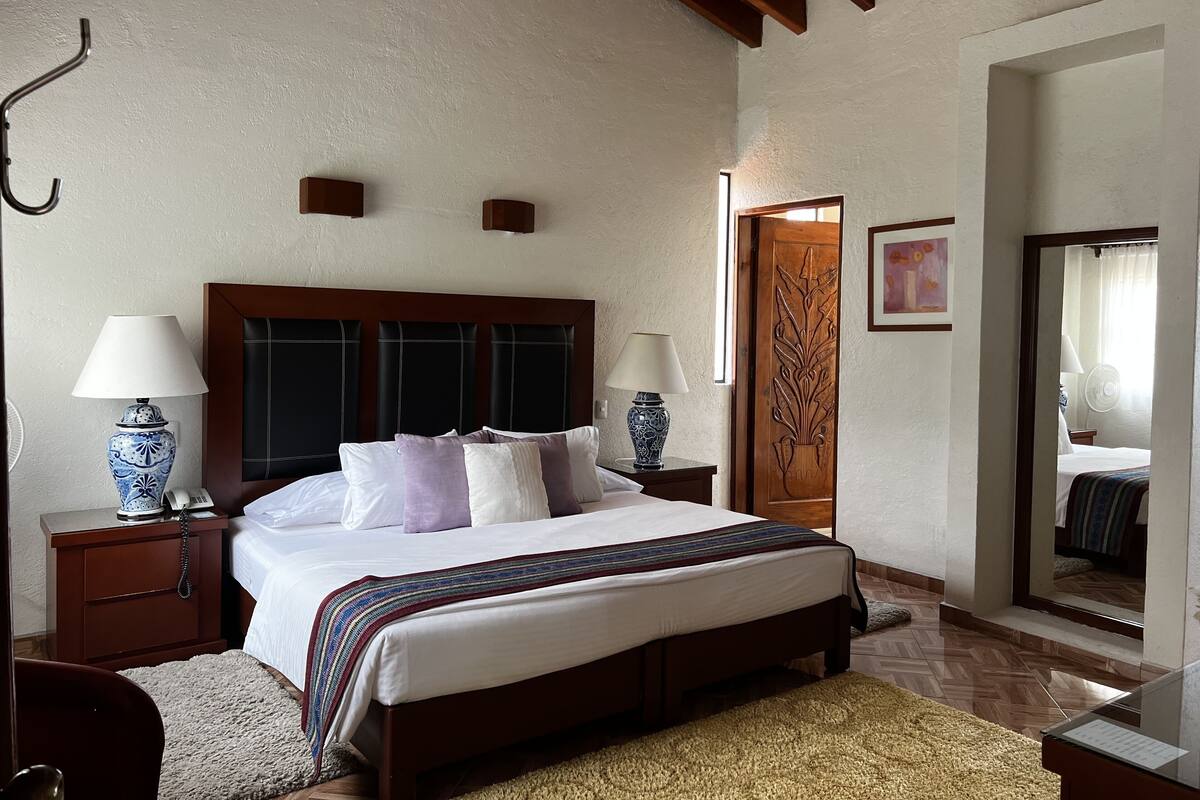 Habitación de hotel con una cama grande, dos mesitas de noche con lámparas, un cabecero de madera, un espejo y una puerta con panel decorativo.
