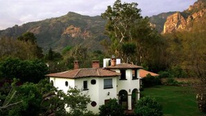 A white building with a tiled roof, surrounded by lush greenery and mountains in the background. - Casa Bugambilia (Tepoztlán)