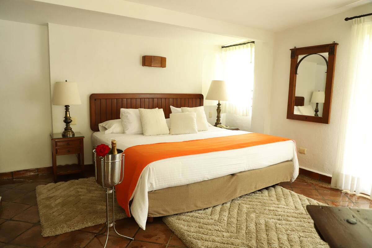 Habitación de hotel ordenada con una cama grande, dos mesitas de noche con lámparas, un espejo y un cabecero de madera.