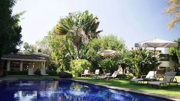 A swimming pool with lounge chairs and umbrellas, surrounded by lush greenery and a building with a tiled roof. - Casa Bugambilia (Tepoztlán)