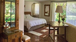 A bedroom with a bed, a wooden nightstand, a table with a lamp, a mirror, and a view of greenery outside. - Casa Bugambilia (Tepoztlán)