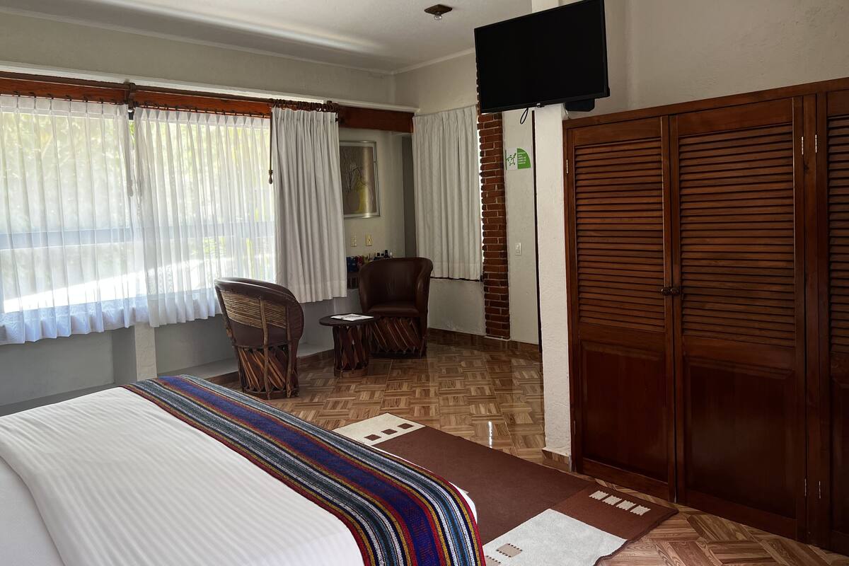Habitación de hotel con cama, armario de madera, televisor, una silla, una mesita y una ventana con cortinas.
