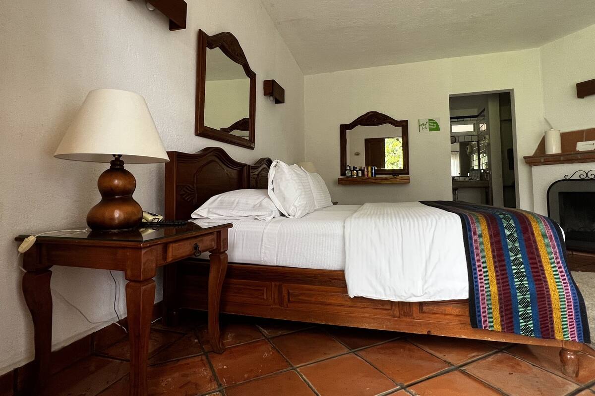 Una cama bien hecha con sábanas blancas, una mesita de noche de madera con una lámpara, un espejo sobre la cama y una manta de colores.
