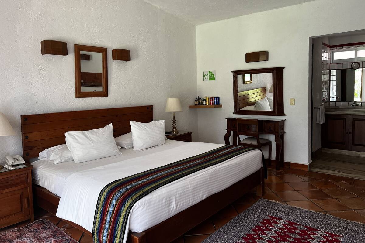 Habitación de hotel con una cama de madera, una mesita de noche con lámpara, un aparador con espejo y un estante con artículos de aseo.