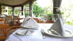 A table set for dining with white napkins, plates, and cutlery, under a covered outdoor area with wooden chairs and a view of greenery. - Casa Bugambilia (Tepoztlán)