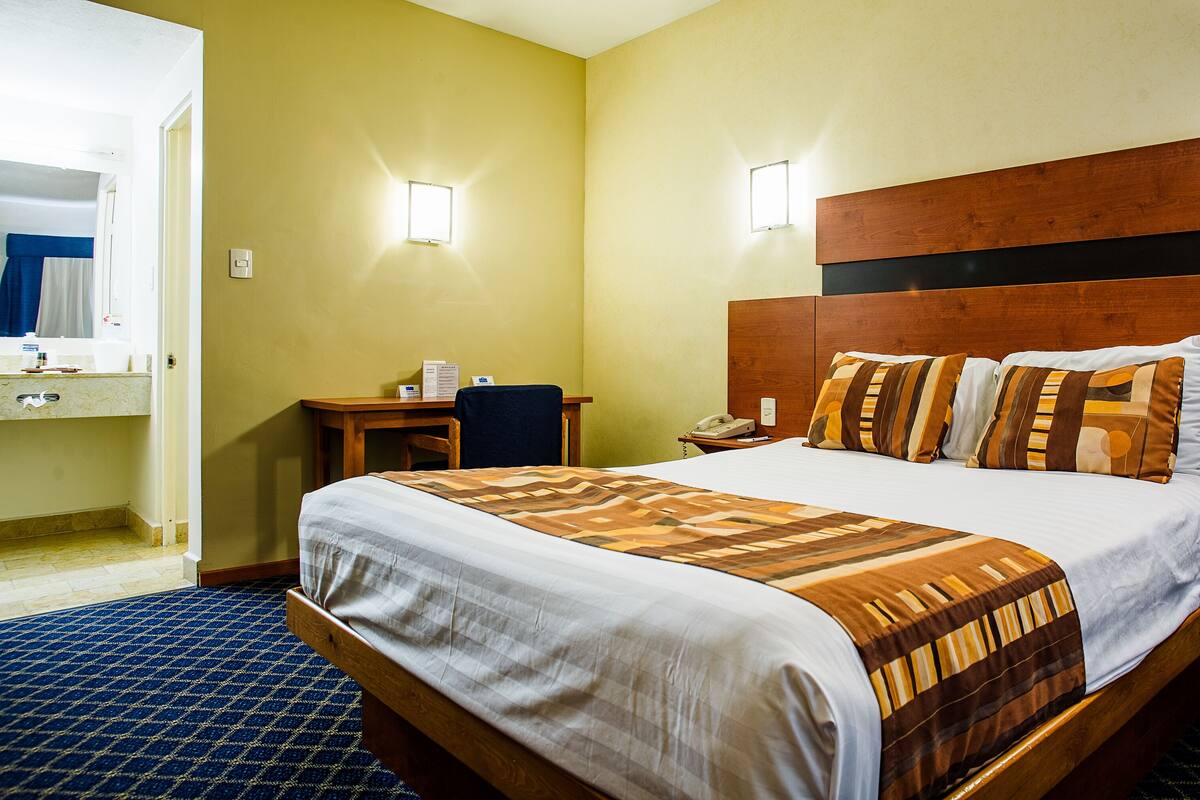 A hotel room with a bed, bedside table, lamp, and a small desk with a chair.