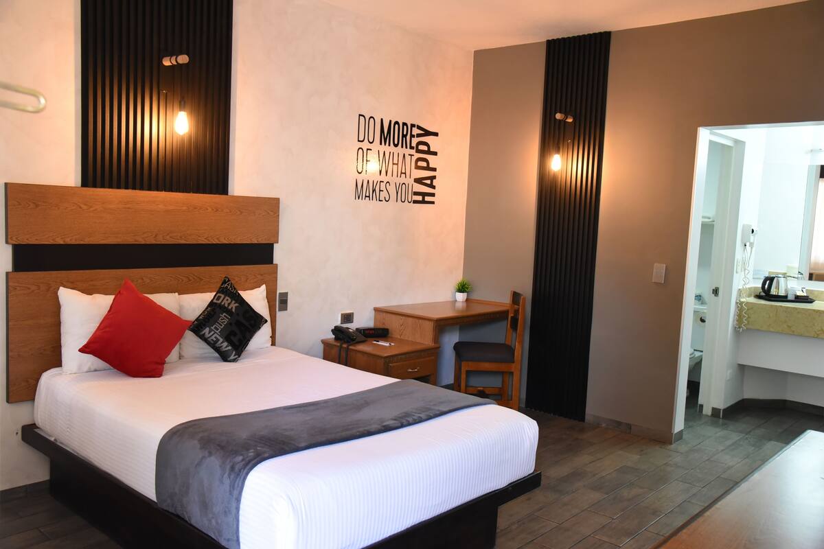 A modern hotel room with a large bed, a desk, and a chair. There is a motivational quote on the wall and a small plant on the desk.