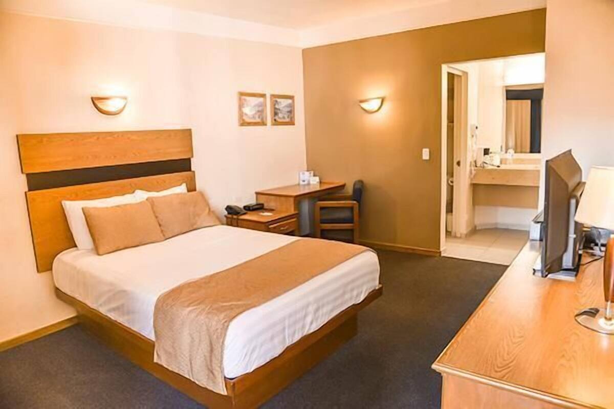 A hotel room with a bed, bedside table, lamp, and a desk with a chair.