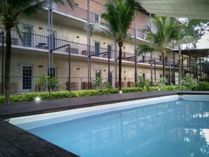A swimming pool with a tiled deck, surrounded by palm trees and a multi-story building with balconies. - NY TH Hotel (Si Racha)
