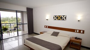 Habitación de hotel con una cama grande, cabecera de madera, mesita de noche y vistas a un balcón con una silla y plantas.