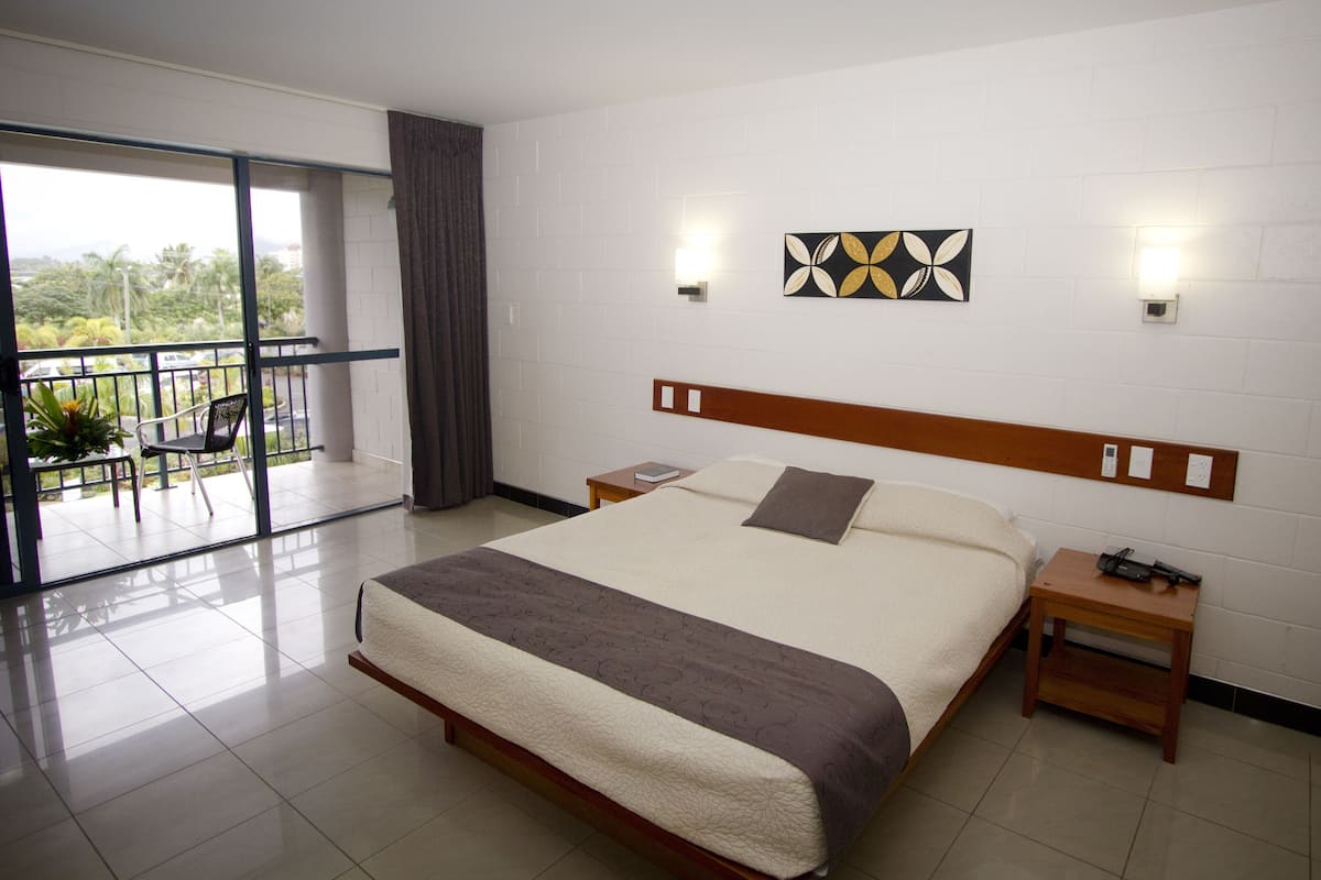A hotel room with a large bed, a wooden headboard, a bedside table, a nightstand, and a view of a balcony with a chair and plants.