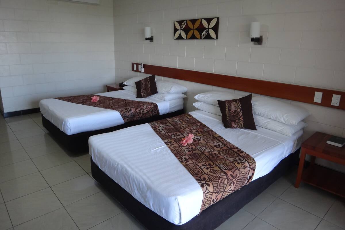 A hotel room with two beds, white bedding, patterned bedspreads, a wooden headboard, a wall-mounted air conditioner, and a decorative wall piece.