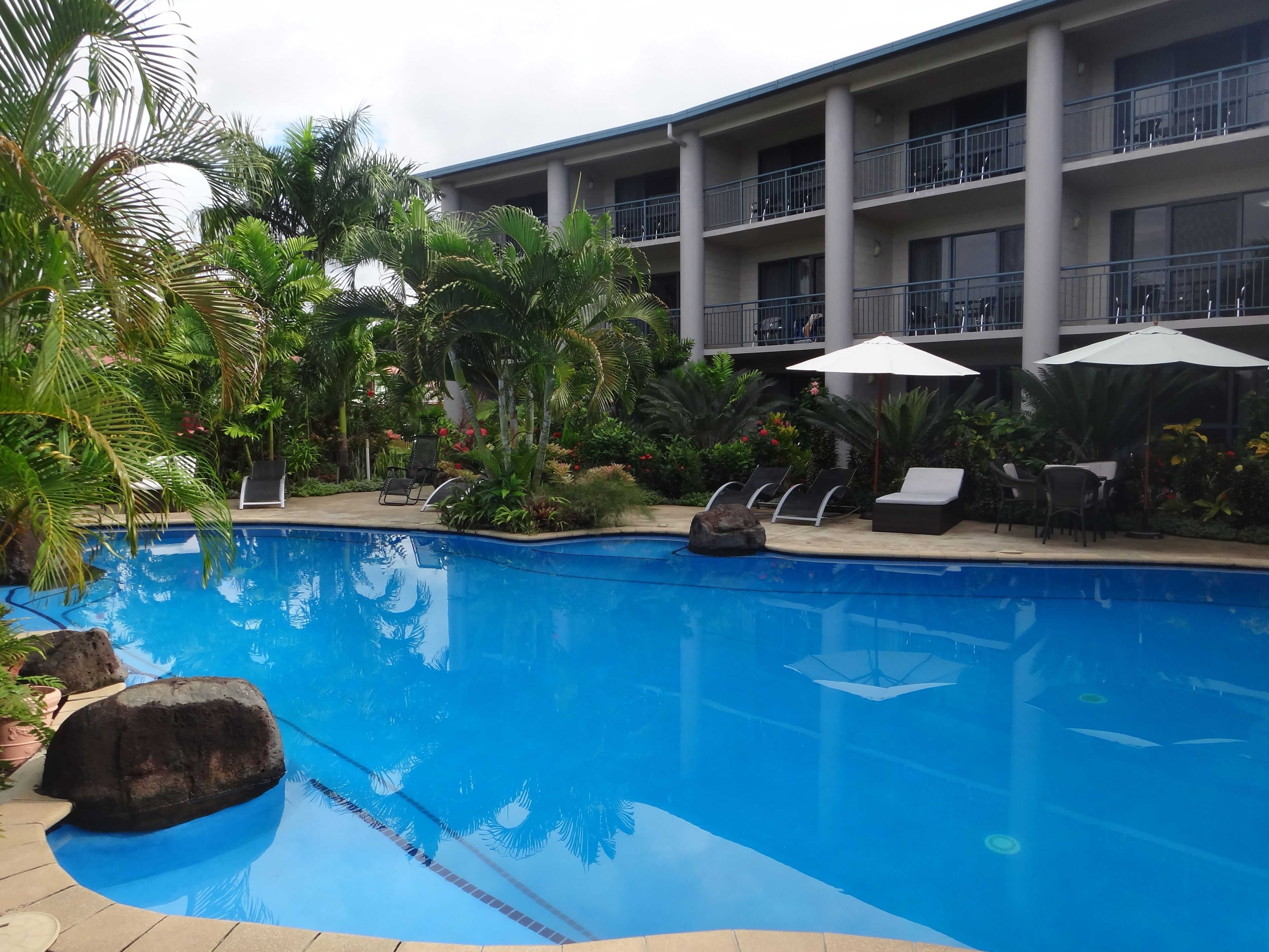 A swimming pool with lounge chairs and umbrellas, surrounded by palm trees and a multi-story building with balconies.