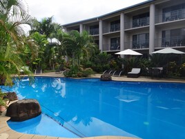 A swimming pool with lounge chairs and umbrellas, surrounded by palm trees and a multi-story building with balconies.