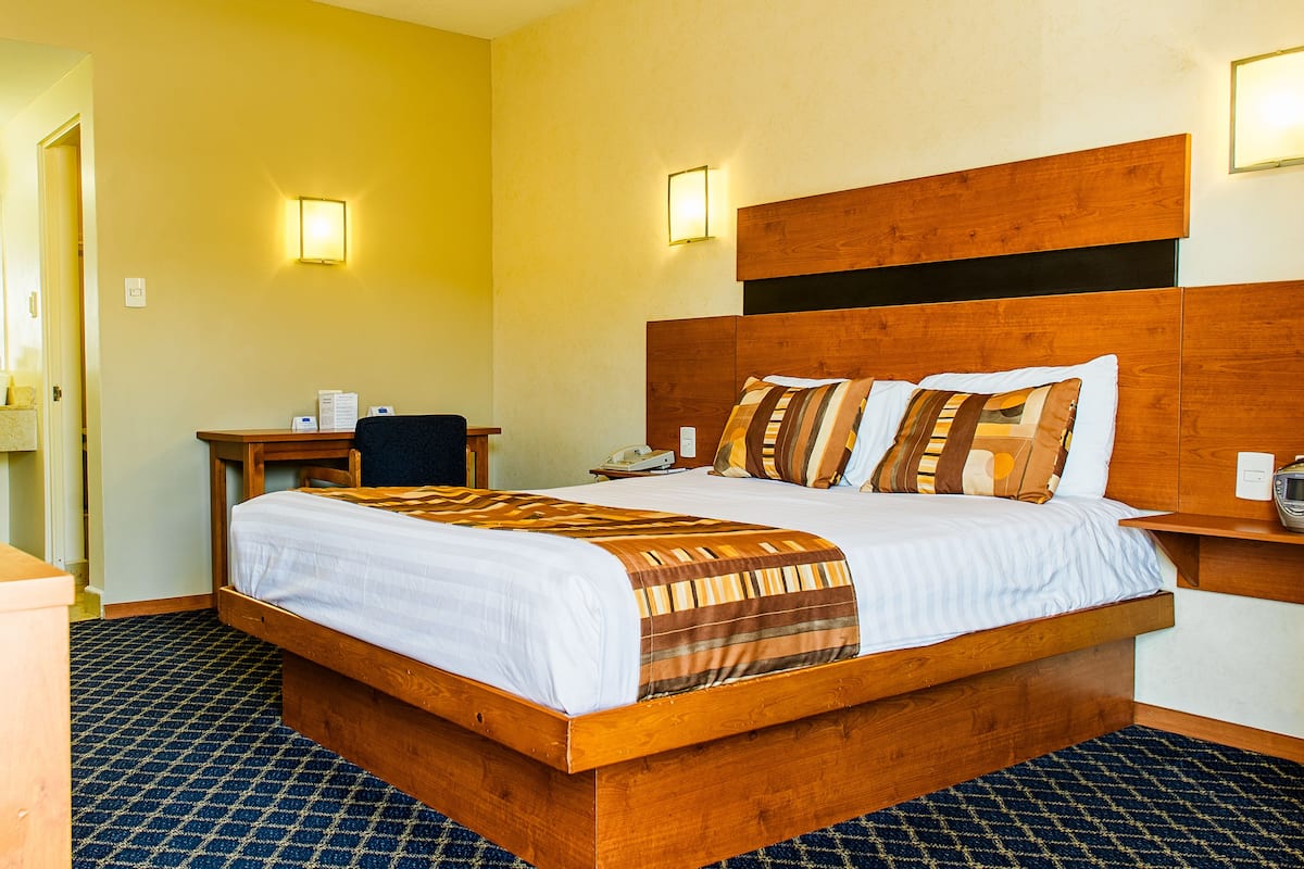 A hotel room with a large bed, a desk, a chair, and a bedside table with a phone and a charger.