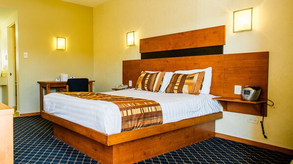 A hotel room with a large bed, a desk, a chair, and a bedside table with a phone and a charger.