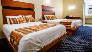 A hotel room with two beds, each with a wooden headboard and patterned bedding. There is a small table with a chair and a nightstand with a lamp. - American Inn Hotel & Suites Delicias (Delicias)