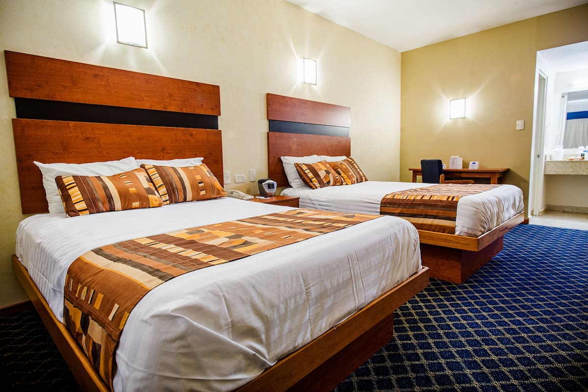 A hotel room with two beds, each with a wooden headboard and patterned bedding. There is a small table with a chair and a nightstand with a lamp.