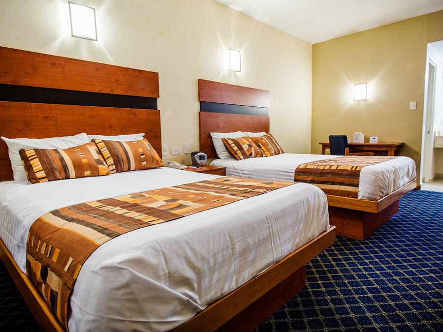 Habitación de hotel con dos camas, cada una con cabecero de madera y ropa de cama estampada. Hay una mesita pequeña con una silla y una mesita de noche con una lámpara.