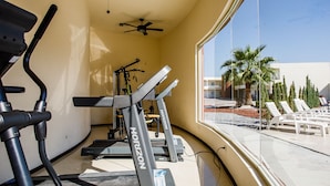 A gym with treadmills and exercise equipment, a ceiling fan, and a view of a pool area with lounge chairs and palm trees. - American Inn Hotel & Suites Delicias (Delicias)