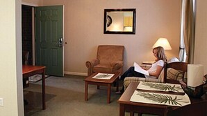 Una habitación con una puerta verde, un sillón marrón, una mesa de madera con un mantel individual, y una persona leyendo un libro.