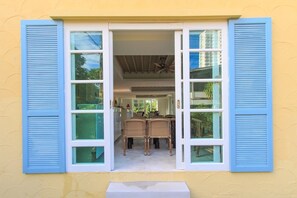 Se ve una habitación con una zona de comedor a través de una ventana con contraventanas azules.