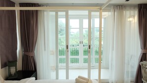 A hotel room with a bed, a small table, and a view of a balcony with greenery. - De Pastel Hotel (Hua Hin)