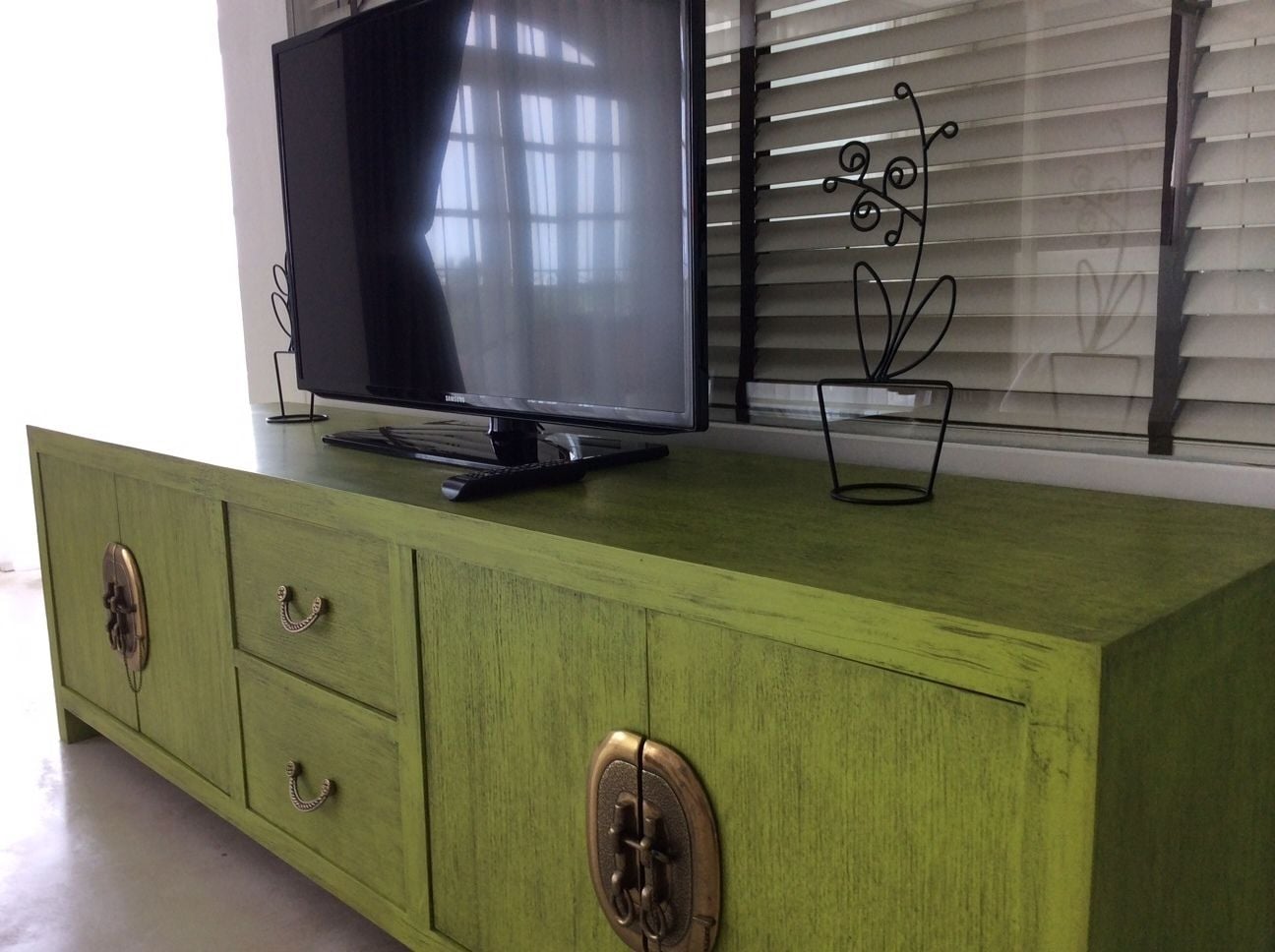 A green cabinet with a flat-screen TV on top, brass handles, and a decorative metal piece on the wall behind.