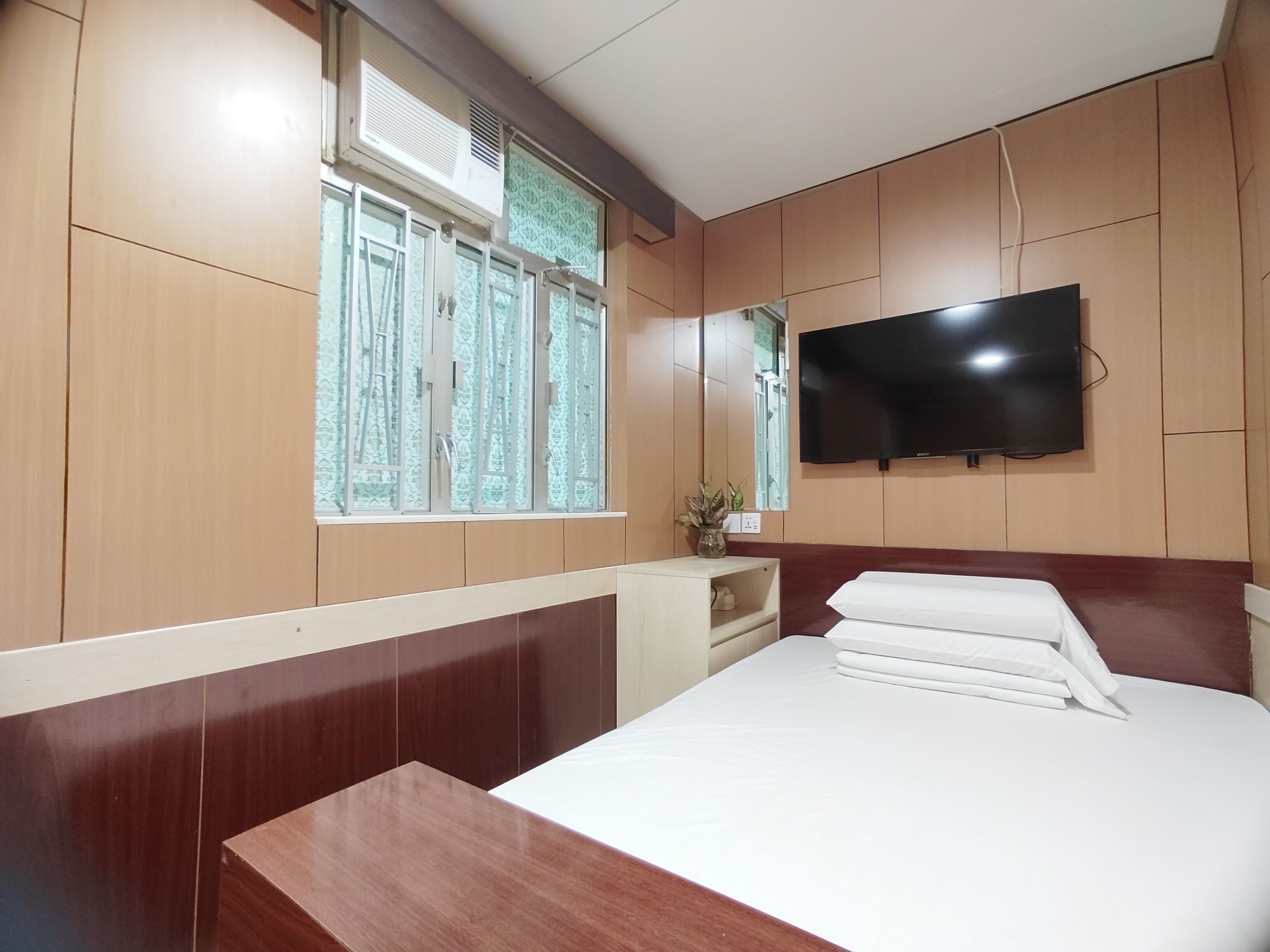 A hotel room with a bed, a TV mounted on the wall, a window with patterned curtains, and a small bedside table.