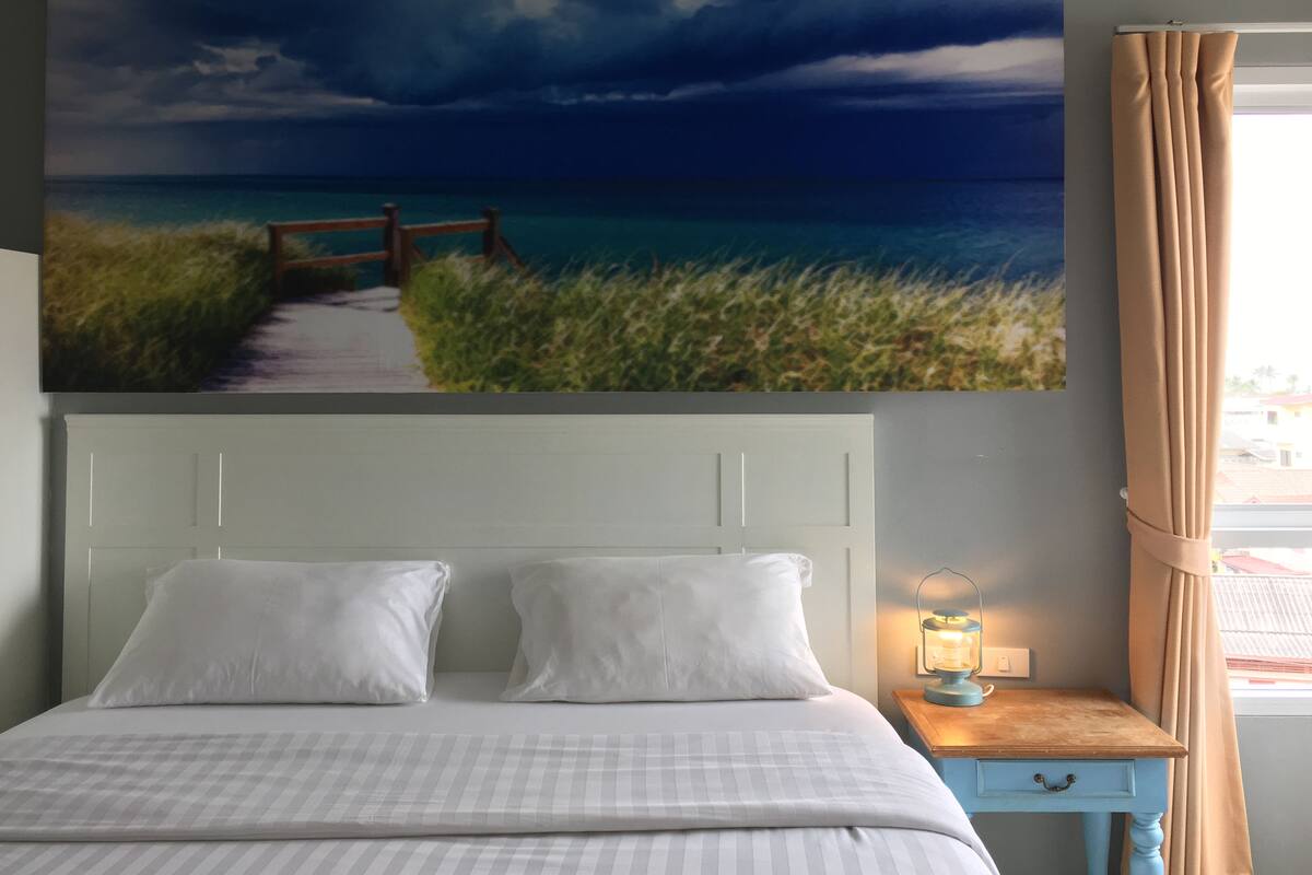 A bedroom with a bed, pillows, a nightstand with a lamp, a painting of a beach scene, and a window with curtains.
