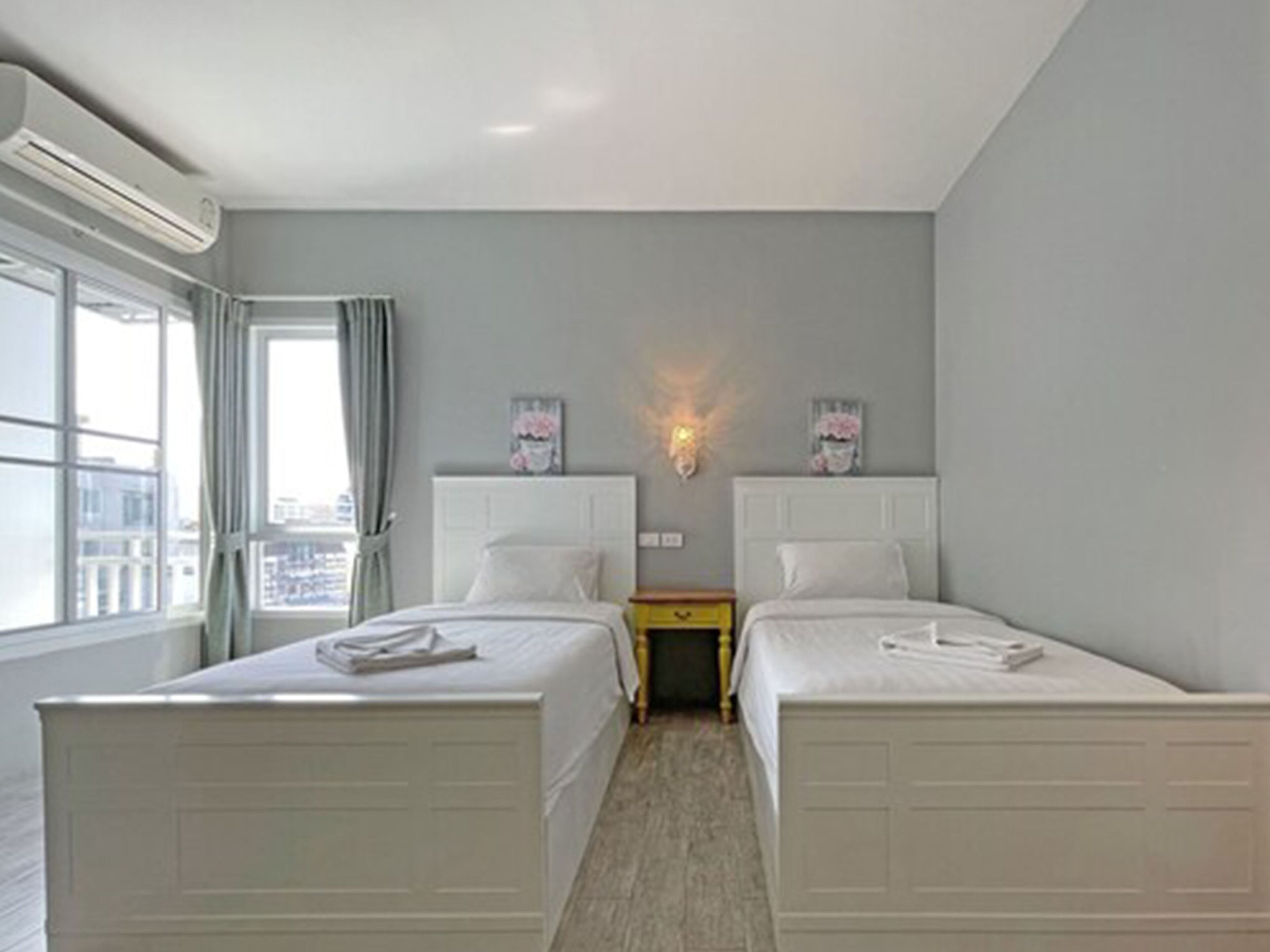 A hotel room with two beds, a window with curtains, an air conditioning unit, and a wall-mounted light fixture.