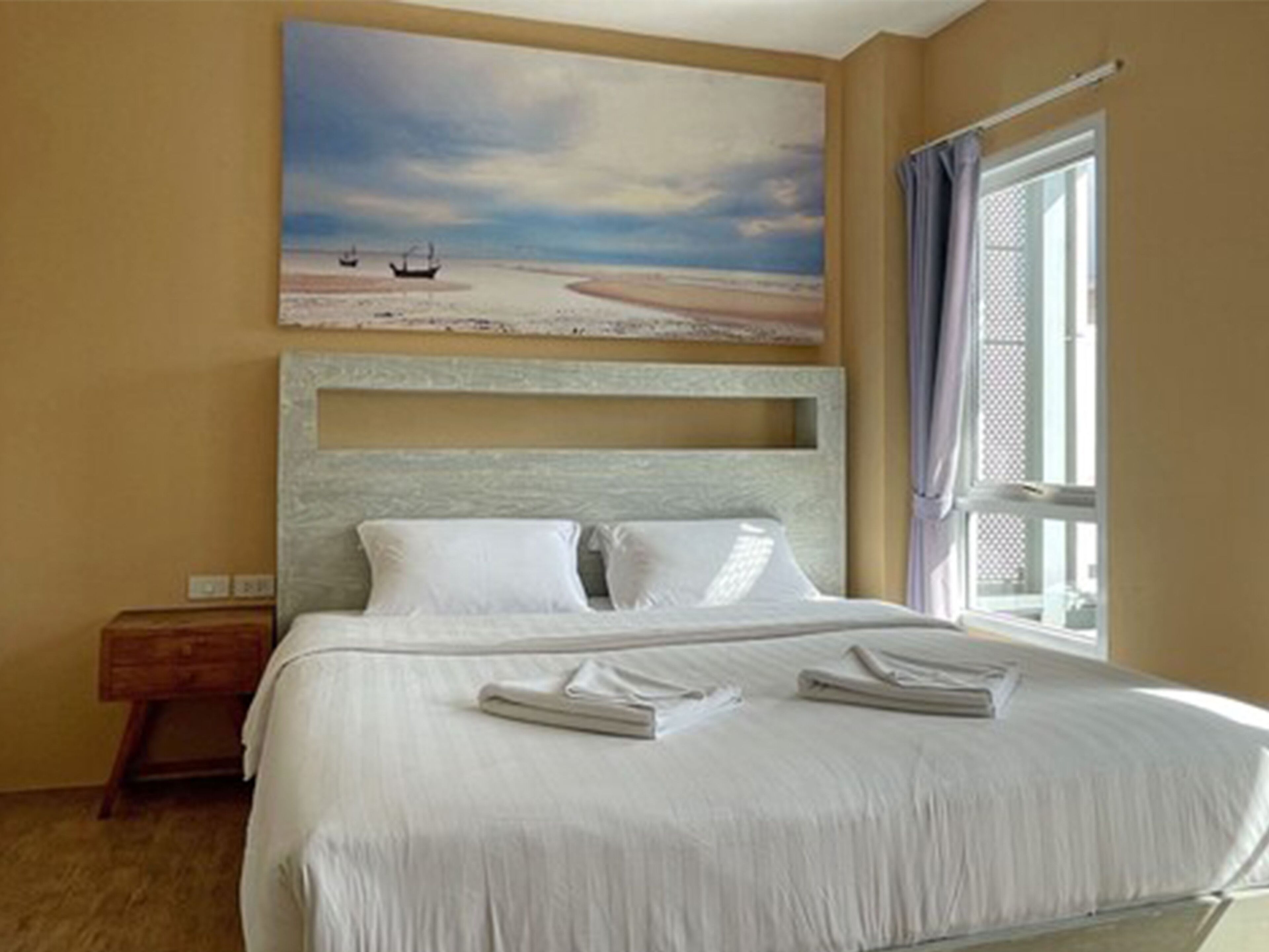 A hotel room with a bed, a nightstand, a painting of a beach scene, and a window with curtains.