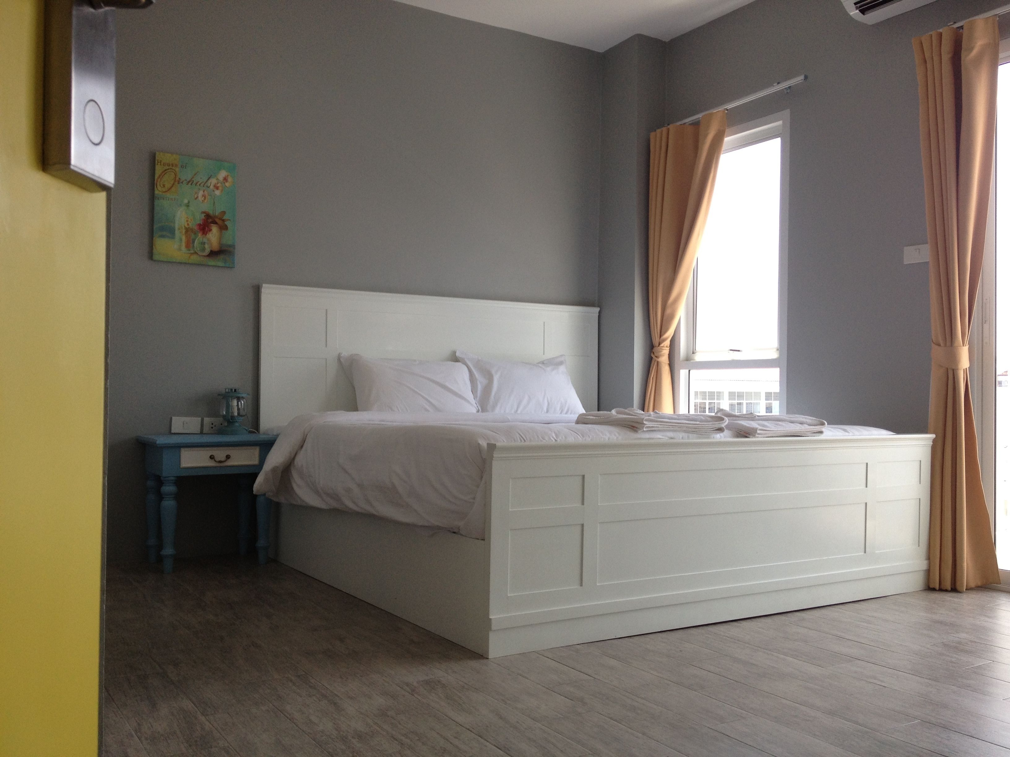A bedroom with a white bed, a nightstand, a small table with a lamp, and a window with curtains.