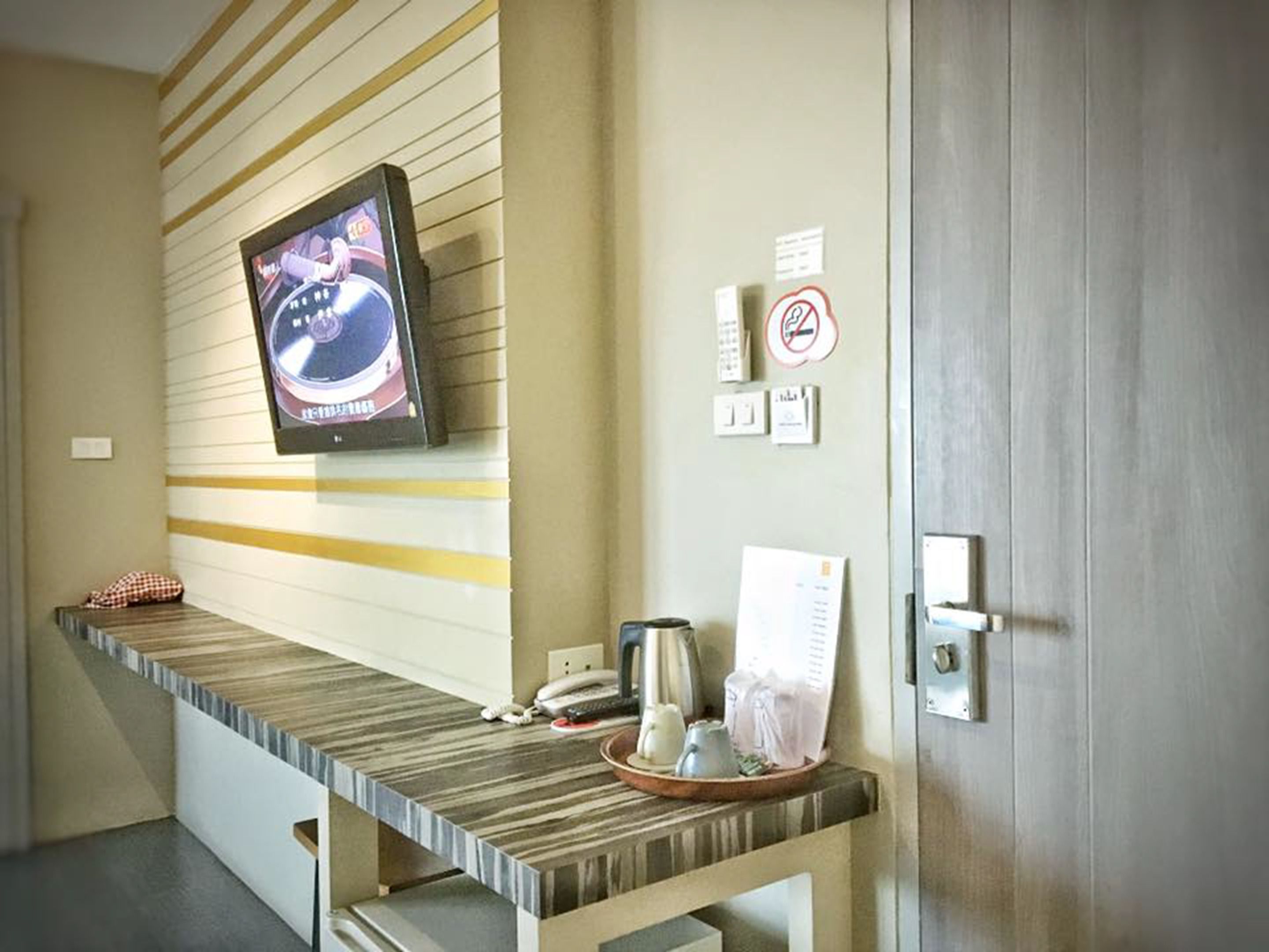 Een hotel receptie met een aan de muur bevestigde televisie, een receptiebalie met een telefoon en een dienblad met spullen, en een deur met een 'niet roken' bord.